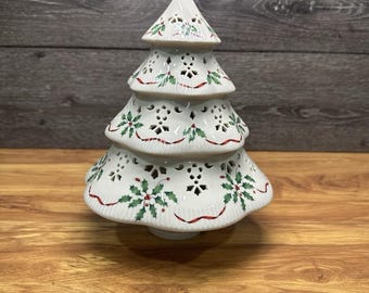 May include: A white ceramic Christmas tree ornament featuring a tiered design with intricate cut-out patterns. The surface is decorated with painted green holly leaves, red berries, and thin red ribbons winding around each of the four levels.