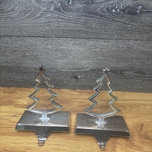 Puede incluir: Dos sujetadores de calcetines navideños plateados. Cada soporte presenta un diseño estilizado de árbol de Navidad con una estrella en la parte superior. Los soportes están hechos de metal brillante y tienen un gancho para colgar calcetines. Los soportes están sobre una superficie de madera.