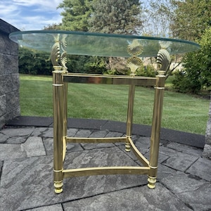 Puede incluir: Una mesa auxiliar dorada con una tapa de cristal transparente. La mesa tiene un marco rectangular con patas curvas y adornos en forma de concha. La mesa mide aproximadamente 61 cm de alto y 51 cm de ancho.