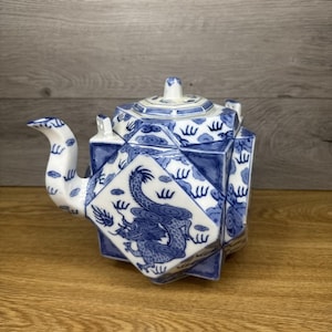 Puede incluir: Una tetera de cerámica azul y blanca con un diseño de dragón. La tetera tiene una forma geométrica con tapa y boquilla curva. El dragón está representado en azul sobre un fondo blanco. La tetera está colocada sobre una superficie de madera.