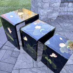 Puede incluir: Juego de tres mesas nido de laca negra. Cada mesa presenta un diseño curvo y está decorada con un motivo de flor de loto en oro, blanco y verde. Las mesas están dispuestas sobre un patio de piedra.