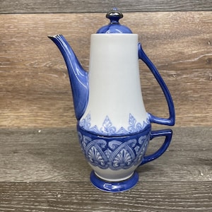 May include: A white and blue ceramic coffee pot with a lid and handle. The pot has a blue spout and handle, with blue decorative patterns around the base and top. The lid is blue with a silver knob.