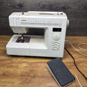 Op de afbeelding: Een witte Brother Compal Opus naaimachine met een zwart voetpedaal. De machine heeft een digitaal display en verschillende steekopties. De machine staat op een houten oppervlak.