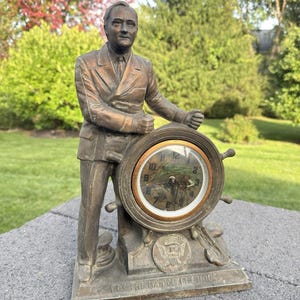 May include: A bronze-toned metal statue of a man in a suit standing beside a ship's wheel clock. The base features the text FOR THE MAN OF THE HOUR and a seal. The clock face displays numbers and a graphic of a ship at sea.