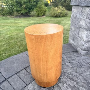 May include: A cylindrical wooden stool with a light brown finish. The stool has a flat top and a slightly wider base. It is approximately 46 cm tall and sits on a gray stone patio.
