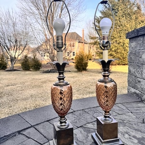 Puede incluir: Dos lámparas de mesa vintage con un cuerpo de vidrio en forma de piña de color cobre y una base cuadrada. Las lámparas tienen un acento floral decorativo y un soporte de pantalla de metal. Las lámparas están iluminadas con bombillas blancas.