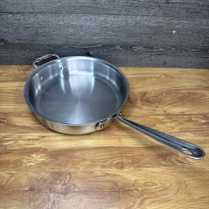 Pode incluir: Uma frigideira de aço inoxidável com uma alça longa e uma alça de laço menor. A panela é redonda com uma superfície brilhante e reflexiva. A panela está em uma superfície de madeira.