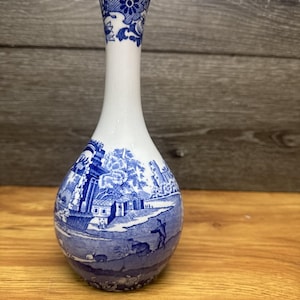 以下が含まれることがあります： 詳細な風景画が描かれた白と青のセラミック花瓶。花瓶は球根状の底と長く細い首を持っています。青いデザインは建物、木、人物を描いています。花瓶の上部には花の模様があります。