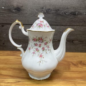 May include: White porcelain teapot with a lid, decorated with pink rose floral patterns and gold trim. The teapot has a curved handle and a spout. The lid is topped with a decorative finial. The teapot is on a wooden surface.