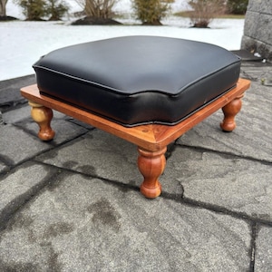 May include: A small, square footstool with a black leather cushion and a wooden base. The base is a warm brown color with turned legs. The footstool is likely used for resting feet or as a decorative accent piece.