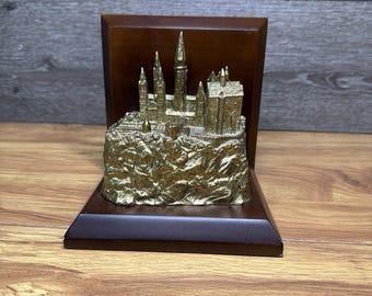 May include: A gold-toned metal sculpture of a castle with multiple towers and spires, mounted on a dark brown wooden L-shaped base. The castle structure sits atop a textured, rocky base, creating a detailed miniature architectural display.