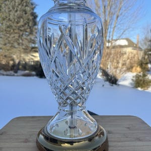 Puede incluir: Una lámpara de mesa de cristal transparente con un patrón de corte de diamante. La lámpara tiene una base dorada y una parte superior plateada. La lámpara está sobre una superficie de madera, con un fondo nevado.