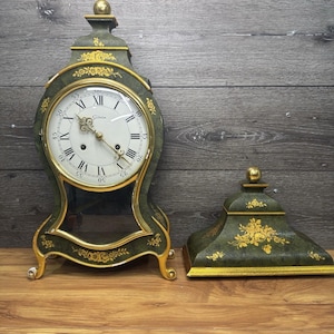 Puede incluir: Un reloj antiguo verde y dorado con una tapa decorativa a juego. El reloj presenta una esfera blanca con números romanos negros y agujas doradas. Diseños florales adornan el marco y la tapa. El reloj se exhibe sobre una superficie de madera.