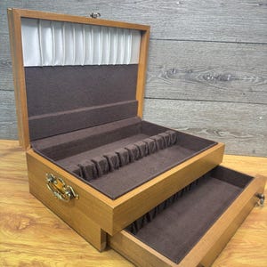 May include: A wooden flatware chest with a hinged lid and a pull-out drawer. The interior is lined with dark brown felt, featuring slotted compartments for silverware. The lid interior has white pleated fabric, and the exterior has gold hardware.