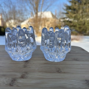 May include: Two clear glass candle holders with a textured, floral-like design. Each holder has a series of vertical, rounded protrusions around the top. The candle holders are sitting on a wooden surface, with a snowy background.