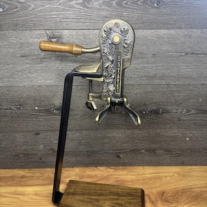 May include: A vintage bronze and black wine bottle opener with a wooden handle and base. The opener features grape vine detailing and the word "ESTATE" on the front. The opener is mounted on a black metal stand.