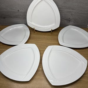 May include: A set of five white, triangular-shaped serving plates. Each plate features a raised, curved design along the edges. The plates are arranged on a wooden surface, with one plate elevated on a gold-colored stand.