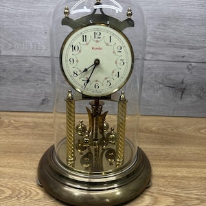 May include: A vintage Kundo clock encased in a clear glass dome. The clock has a cream-colored face with black numerals and hands. The base and decorative elements are gold-toned. The clock features a pendulum and is set on a round base.