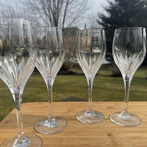 Op de afbeelding: Vier heldere kristallen wijnglazen met een decoratief geëtst bladerontwerp. De glazen staan op een houten oppervlak, met een vage buitenachtergrond. De glazen hebben een lange, slanke steel en een brede kelk.