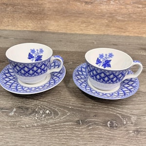 May include: Two matching teacups and saucers. The white teacups have blue floral designs inside and a blue diamond pattern on the outside. The saucers have a matching blue diamond pattern. The teacups and saucers are on a wooden surface.
