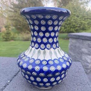 Puede incluir: Un jarrón de cerámica azul y blanco con la parte superior acampanada y la base redondeada. El jarrón está decorado con un patrón repetitivo de círculos azules con detalles en blanco y rojo. También hay formas verdes en forma de hoja.