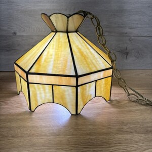 Könnte beinhalten: Eine Vintage-Hängeleuchte aus Buntglas. Der Lampenschirm hat eine sechseckige Form mit einem gewellten oberen und unteren Rand. Das Glas ist gelb und weiß mit schwarzen Linien. Eine Messingkette ist oben befestigt.