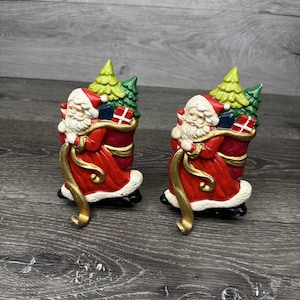May include: Two Santa Claus figurines, each with a red coat, white beard, and hat, carrying a sack of gifts and a Christmas tree. The figurines have gold accents and black bases, perfect for holiday decor.