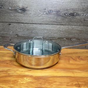 May include: A stainless steel sauté pan with a polished finish, featuring a long handle, a smaller helper handle on the opposite side, and a clear glass lid with a metal handle. The pan has a wide, flat base and straight vertical sides.