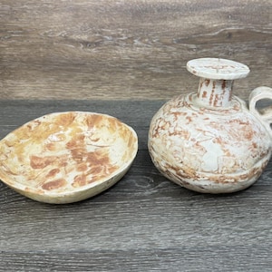 Puede incluir: Un cuenco de cerámica y una jarra pequeña con asa. El cuenco es de forma ovalada con un patrón jaspeado marrón claro y beige. La jarra es redonda con una parte superior plana y un asa. Ambos artículos tienen una base texturizada blanquecina con acentos marrones.