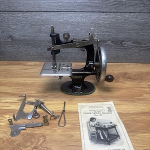 Puede incluir: Una máquina de coser Singer modelo 20 negra vintage con manivela y placa de aguja plateadas. Se incluyen varios accesorios de costura metálicos sueltos y un manual de instrucciones titulado Instructions for using Singer Sewing Machine No. 20.
