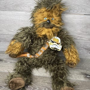 May include: A plush Chewbacca doll from Star Wars, featuring brown and tan fur, blue eyes, and a bandolier. The doll has a Star Wars tag attached. The plush toy is sitting on a wooden surface.