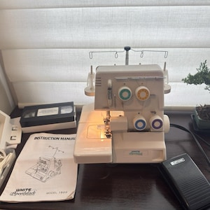 Könnte beinhalten: Eine weiße White Speedylock Modell 1500 Overlock-Nähmaschine mit vier farbigen Spannungsreglern, einem schwarzen Fußpedal, einer Bedienungsanleitung, einem Kunststoff-Zubehörfach mit Spulen und einer schwarzen VHS-Kassettenhülle auf einer dunklen Fläche.