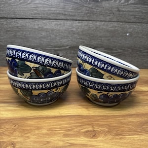 Könnte beinhalten: Drei Keramikschalen mit einem handgemalten Muster aus dunkelblauen Trauben und grünen Blättern auf cremefarbenem Grund. Jede Schale hat einen dekorativen dunkelblau-weißen geometrischen Rand entlang der Öffnung und eine abgerundete Form.