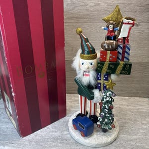 May include: A decorative Christmas nutcracker figurine with a white beard, green jacket, and red and white striped pants. The nutcracker holds a small Christmas tree and stands on a snowy base with a blue toy train. A box with red stripes is on the left.