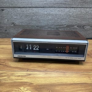 Puede incluir: Un radio reloj digital AM-FM Soundesign rectangular con una parte superior de imitación madera y un panel frontal plateado. El dispositivo cuenta con una pantalla negra que muestra la hora 11:22 PM, un sintonizador de radio y etiquetas.