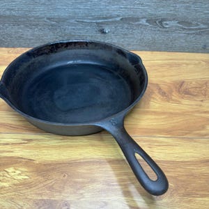 以下が含まれることがあります： 黒色の鋳鉄製フライパン。長いハンドルと丸みを帯びた調理面があります。木製の背景に置かれ、そのクラシックなデザインと調理の可能性を示しています。