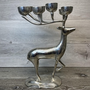 Könnte beinhalten: Ein silberfarbener kerzenhalter in Hirschform mit fünf Kerzenschalen. Der Hirsch steht auf einem runden Sockel mit erhobenem Kopf. Die Kerzenschalen sind auf dem Geweih des Hirsches angeordnet.
