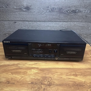 Puede incluir: Una pletina de casete doble Sony de color negro con dos compartimentos etiquetados como Deck A y Deck B, ambos con función de auto-reverse. El panel frontal incluye una pantalla digital central, varios botones de control, un mando de balance, un mando de nivel de grabación y una toma de auriculares.