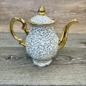 May include: A vintage white porcelain teapot with a gold floral pattern and gold accents. The teapot features a curved handle and spout, and a lid with a gold finial. The teapot is a decorative item.