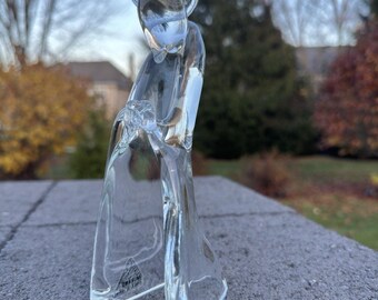 May include: A clear glass angel figurine with a halo. The angel is depicted in a flowing gown, with a serene expression. The figurine is standing on a gray surface, with a blurred outdoor background.