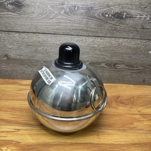 May include: A spherical stainless steel container with a polished finish, featuring a black knob on top and a metal ring handle on the side. A white rectangular sticker is attached to the surface with the text "SEE WARNING ON BOTTOM BEFORE USE".