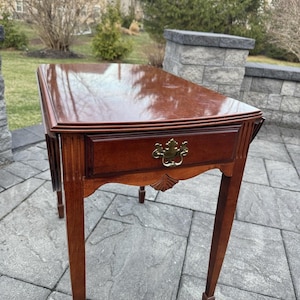 May include: A mahogany-stained wooden side table with a single drawer and brass hardware. The table has a rectangular top with a curved front edge and slender, tapered legs. The drawer features a decorative pull.