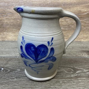 May include: A stoneware pitcher with a light gray base and a curved handle. The pitcher is decorated with a blue heart-shaped floral design. The pitcher has a slightly textured surface and a rounded base.