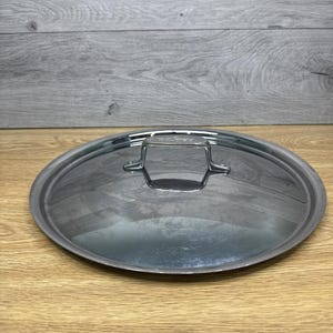 May include: A stainless steel pot lid with a handle. The lid is round and has a brushed metal finish. The handle is a small, rectangular shape. The lid is sitting on a wooden surface.