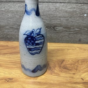Può includere: Una bottiglia in gres con finitura screziata grigio chiaro e collo stretto. La bottiglia presenta un design a mela blu e dettagli a onda blu. La bottiglia è esposta su una superficie di legno.
