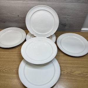 Può includere: Un set di piatti in ceramica bianca di varie dimensioni. I piatti presentano un design semplice ed elegante con un bordo leggermente rialzato. Sono disposti su una superficie di legno, evidenziando la loro estetica pulita e minimalista.