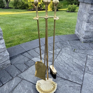Op de afbeelding: Een messing haardstel bestaande uit een standaard met een schelpvormige voet, een schep en een borstel. Elk van de drie gereedschappen met lange steel is voorzien van een decoratieve, schelpvormige knop van gepolijst goudkleurig metaal.