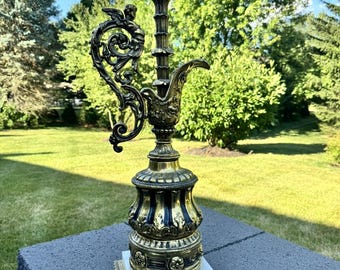May include: An ornate brass and black table lamp with a white marble base. The lamp features intricate detailing, including a decorative handle with an angel figure. The lamp is set outdoors on a gray stone surface, with a green lawn and trees in the background.