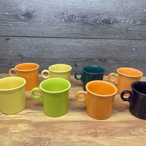 Op de afbeelding: Een verzameling van zeven keramische mokken in diverse effen kleuren, waaronder felgeel, limoengroen, oranje, blauwgroen en donkerpaars. Elke mok heeft een gladde, glanzende afwerking, een afgerond lichaam en een cirkelvormig handvat.