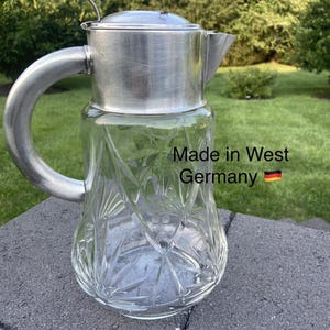 May include: A vintage glass and metal pitcher with a decorative etched design. The pitcher has a silver-colored metal lid, handle, and spout. The text "Made in West Germany" is visible on the glass.